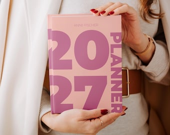 Könnte beinhalten: Ein Hardcover-Planer mit hellrosa Einband, der den großen violetten Text 2027 und das vertikal auf der rechten Seite gedruckte Wort PLANNER zeigt. Der Name ANNE FISCHER ist in kleiner, dünner, heller Schrift oben mittig aufgedruckt.
