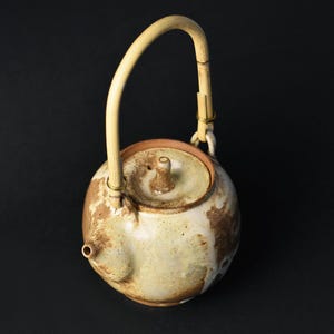 Op de afbeelding: Een crèmekleurige keramische theepot met bruine en bruingele gevlekte patronen. Het heeft een ronde body, een kleine tuit en een deksel met een knop. De handgreep is van bamboe en is bevestigd met metalen bevestigingen. Traditioneel ontwerp.