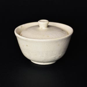 Può includere: Una piccola ciotola in ceramica bianco sporco con coperchio abbinato. La ciotola ha una forma arrotondata e una superficie leggermente ruvida. Il coperchio presenta una piccola maniglia arrotondata. La ciotola e il coperchio sono su sfondo nero.