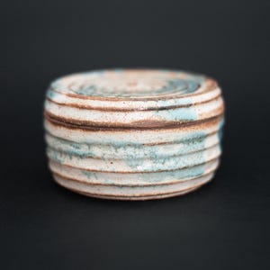 Puede incluir: Un pequeño recipiente de cerámica hecho a mano con tapa. La pieza cilíndrica presenta bandas horizontales blancas, marrones y azul claro. La cerámica tiene un aspecto texturizado y orgánico, sobre un fondo negro.