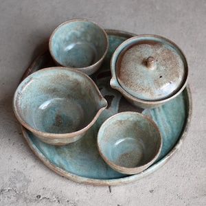 May include: A teal and brown ceramic tea set. The set comprises a teapot with a lid, a serving bowl with a spout, two small bowls, and a round tray. The pottery has a rustic, textured finish. The set is designed for serving tea.