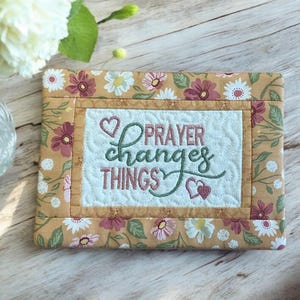May include: A rectangular quilted item with a floral pattern in shades of tan, pink, and white. The center features the embroidered text "PRAYER changes THINGS" in green and pink, with heart accents. The item is likely a decorative piece.