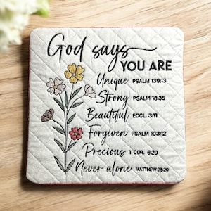 May include: Square, quilted coaster with the text "God says YOU ARE Unique, Strong, Beautiful, Forgiven, Precious, Never alone." A floral design with yellow, pink, and brown flowers is on the left side. The coaster is on a wooden surface.