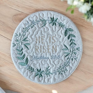 May include: A round, light blue quilted coaster with the embroidered text "HE IS RISEN" and "Matthew 28:6". The coaster is encircled by a green leaf design.