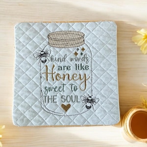 May include: A light blue quilted square coaster with a jar design and the text "kind words are like honey sweet to the soul." The jar has a woven lid, bees, and a heart. Yellow flowers and a small gold-colored pot are also present.