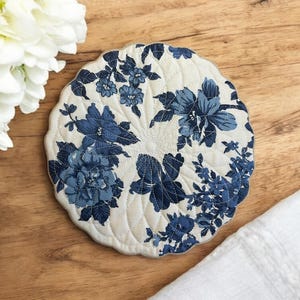 May include: Circular, quilted coaster with a cream backdrop and a blue floral design. The coaster has a scalloped edge and is on a wooden surface. White flowers are in the background.