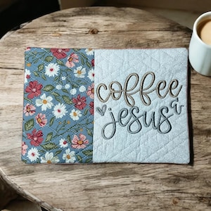 May include: A quilted coaster with the words "coffee jesus" embroidered on a light blue background. The coaster is next to a floral patterned fabric and a white mug filled with coffee.