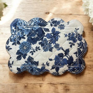 May include: A quilted placemat with a scalloped edge. The placemat has a white background with a pattern of blue flowers and leaves. The edge is trimmed with a blue stitch.