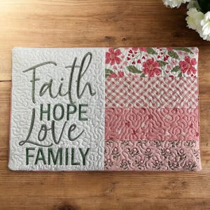 May include: A quilted rectangular decorative piece with the words "Faith, Hope, Love, Family" embroidered in green. The right side features pink floral and patterned fabric sections. The piece is placed on a wooden surface.