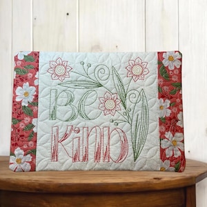 May include: A quilted clutch bag with the words "BE KIND" embroidered in green and red. The central panel is light green with floral embroidery. The sides are red with white floral patterns. The bag is on a wooden table.