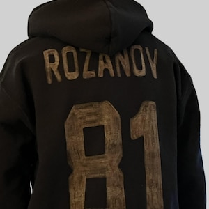 Peut inclure: Sweat à capuche noir avec le nom "ROZANOV" et le numéro "81" en couleur beige. Le sweat à capuche a une capuche et est fait d'une matière douce. Le design est au dos du sweat.