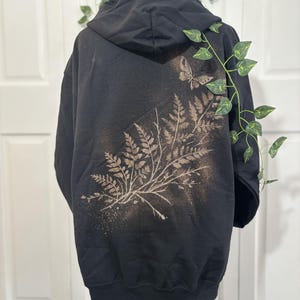 Peut inclure: Un sweat-shirt à capuche noir présentant un motif décoloré au dos. Le graphisme se compose d'une grande fougère s'étendant en diagonale sur le tissu, accompagnée d'une petite silhouette de papillon près de l'épaule supérieure droite.