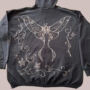 Könnte beinhalten: Ein schwarzer Kapuzenpullover mit einer großen, hellen Grafik auf der Rückseite. Das Design zeigt einen zentralen Nachtfalter mit langen, geschwungenen Flügeln, umgeben von zarten, rankenden Mustern und kleinen floralen Elementen.