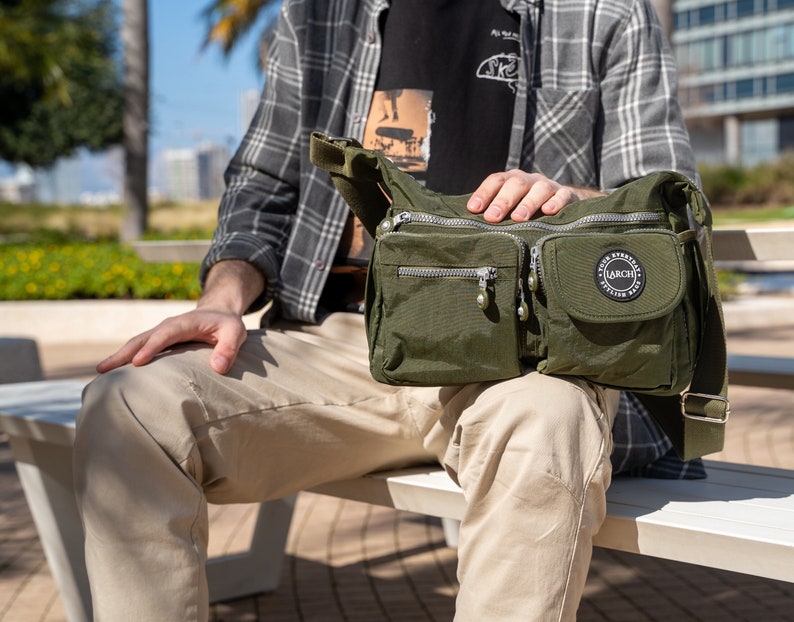 May include: A person wearing a plaid shirt and khaki pants is sitting on a bench. They are carrying a green nylon crossbody bag with a zippered pocket and a circular logo that says "LARCHI" in white letters.