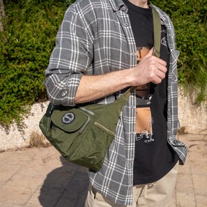 May include: A person wearing a gray and white plaid shirt with a black t-shirt underneath. The t-shirt has a graphic print of a palm tree and the text "Venice". The person is carrying a green crossbody bag with a black zipper and a black circle logo that says "Respect".