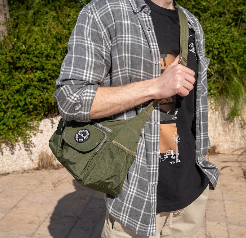 May include: A person wearing a gray and white plaid shirt with a black t-shirt underneath. The t-shirt has a graphic print of a palm tree and the text "Venice". The person is carrying a green crossbody bag with a black zipper and a black circle logo that says "Respect".