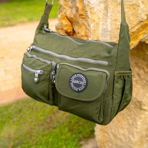 May include: A green nylon shoulder bag with multiple zippered compartments and a large front pocket with a black circular patch that reads "Your Everyday Stylish Bags LARCH".