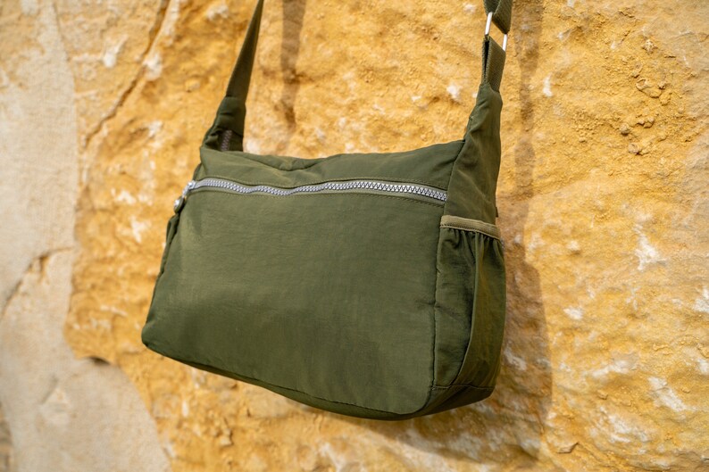 May include: A dark green crossbody bag with a zippered pocket and a small pocket on the side. The bag is made of a soft, lightweight material.