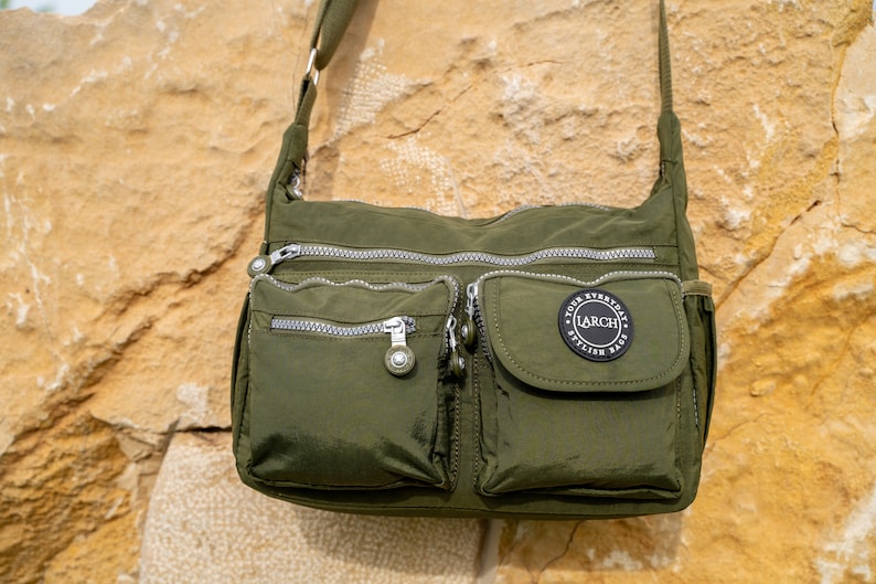 May include: A green nylon crossbody bag with multiple zippered pockets and a black circular patch that reads "YOUR EVERYDAY LARCH STYLISH BAGS".