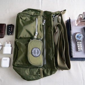 May include: A green canvas shoulder bag with a zippered pocket and a black logo that says "Larch" on the front. The bag is on a white surface with other items, including a brown leather wallet, a car key, a white phone charger, a pair of white earbuds, a black notebook, a silver pen, a silver watch, a black glasses case, and a bottle of perfume.