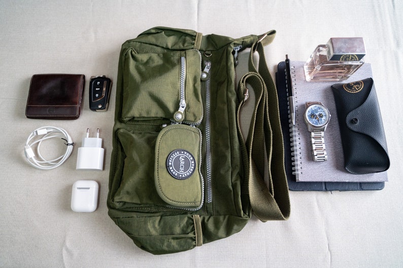 May include: A green canvas shoulder bag with a zippered pocket and a black logo that says "Larch" on the front. The bag is on a white surface with other items, including a brown leather wallet, a car key, a white phone charger, a pair of white earbuds, a black notebook, a silver pen, a silver watch, a black glasses case, and a bottle of perfume.