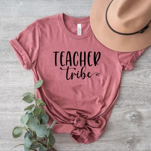 Peut inclure: Un t-shirt mauve avec le texte "Teacher Tribe" en lettres noires. Le t-shirt est noué à l'avant et est posé sur une surface en bois.