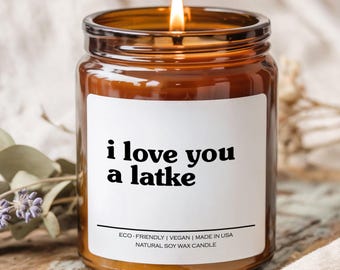 May include: A lit, amber-colored glass jar candle with a white label. The label reads "i love you a latke" in black text. The candle is eco-friendly, vegan, and made in the USA. It is a natural soy wax candle.