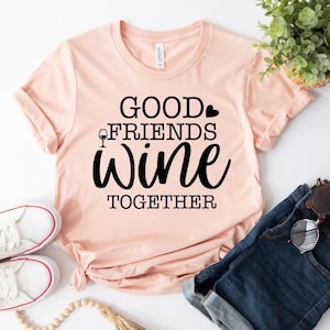 May include: A light pink t-shirt with the words "GOOD FRIENDS WINE TOGETHER" in black script. The shirt has a crew neck and short sleeves. White sneakers and blue jeans are visible.