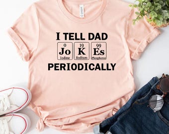 Op de afbeelding: Een roze t-shirt met de tekst "I tell dad jokes periodically" en een afbeelding van drie chemische elementen: Jodium, Natrium en Fosfor.