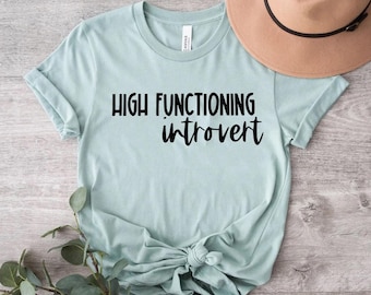 May include: A light blue t-shirt with the text "HIGH FUNCTIONING introvert" in black. The shirt is knotted at the bottom and is next to a brown hat and some greenery. The shirt is made of a soft material.