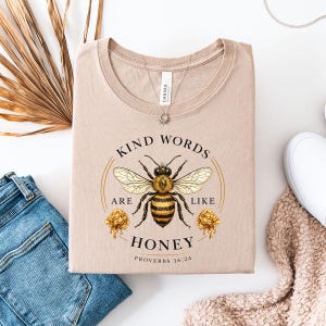 以下が含まれることがあります： 蜂のグラフィックと「Kind Words Are Like Honey Proverbs 16:24」の文字が入ったベージュ色のTシャツ。 蜂は黄色、黒、茶色で、ハニカムが描かれています。 Tシャツは折りたたまれています。