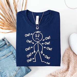 May include: A navy blue t-shirt with a white stick figure design. The stick figure has a sad face and arrows pointing at it with the word "OW!" written on each arrow. A silver necklace with a sun pendant is visible.