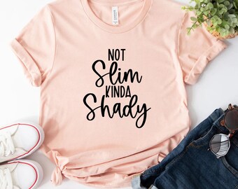 May include: A light peach-colored t-shirt with the words "NOT Slim Kinda Shady" in black script. The shirt is styled with rolled-up sleeves, a knot at the waist, and paired with blue jeans and white sneakers.