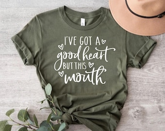 Puede incluir: Una camiseta verde con el texto "I've got a good heart but this mouth" en letras blancas con pequeños corazones alrededor del texto.