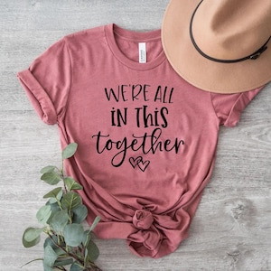 Puede incluir: Una camiseta de color rosa polvoriento con el texto "We're all in this together" en escritura negra con dos pequeños corazones debajo del texto.