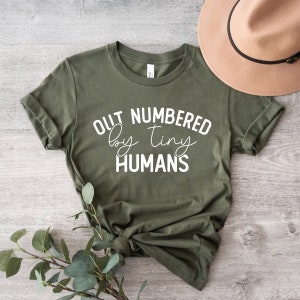 Può includere: Una maglietta verde con testo bianco che dice "Outnumbered by tiny humans".