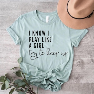 May include: A pale blue t-shirt with the text "I KNOW I PLAY LIKE A GIRL try to keep up" in black. A tan hat is visible in the upper right corner. The shirt is knotted at the bottom and rests on a wooden surface.