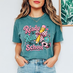 Könnte beinhalten: Ein kurzärmeliges T-Shirt in Petrol mit einem Grafikdruck und dem Text "Ready to Rule the School" in Pink und Petrol. Das Design zeigt einen gelben Bleistift mit einer rosa gepunkteten Schleife und einen petrolfarbenen Rucksack.