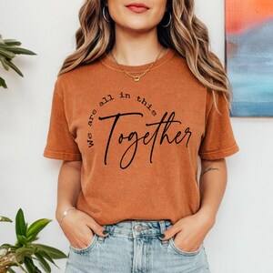 Könnte beinhalten: Ein rostfarbenes Kurzarm-T-Shirt mit dem schwarzen Schriftzug "We are all in this Together" auf der Vorderseite. Das Shirt hat einen Rundhalsausschnitt und eine lockere Passform und wird mit einer hellblauen Jeans aus Denim kombiniert.