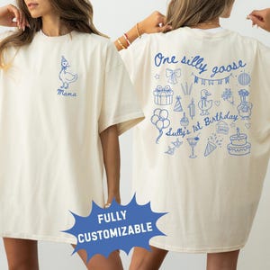 Peut inclure: T-shirt cr&egrave;me avec des graphiques bleus. L'avant pr&eacute;sente une oie portant un chapeau de f&ecirc;te et le mot "Mama". Le dos affiche le texte "One silly goose" et "Sully's 1st Birthday" avec des illustrations festives, dont des ballons et un g&acirc;teau.