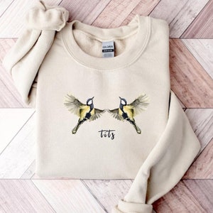 May include: A beige sweatshirt with a graphic of two birds flying with the word "tits" printed below them.