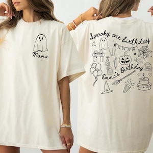 May include: Ivory-colored t-shirt with a black ghost graphic and the word "Mama" on the front. The back features a "Spooky one birthday" design with birthday-themed illustrations, including a pumpkin, cake, and balloons.