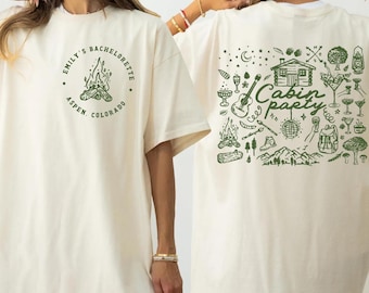 May include: Ivory-colored t-shirt with green text and graphics. The front features "Emily's Bachelorette Aspen, Colorado" around a campfire illustration. The back displays "Cabin Party" with camping-themed icons.