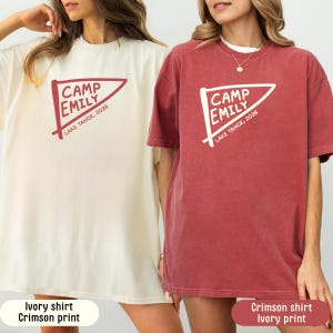 May include: Two short-sleeve crew neck t-shirts. The left shirt is ivory with a crimson graphic of a pennant flag featuring the text CAMP EMILY LAKE TAHOE, 2026. The right shirt is crimson with an ivory graphic of the same pennant flag design.