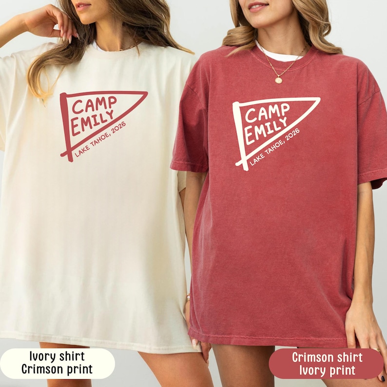 May include: Two short-sleeve crew neck t-shirts. The left shirt is ivory with a crimson graphic of a pennant flag featuring the text CAMP EMILY LAKE TAHOE, 2026. The right shirt is crimson with an ivory graphic of the same pennant flag design.