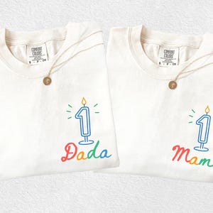 May include: Two off-white t-shirts with the text "Dada" and "Mama" in rainbow colors, each featuring a number one candle graphic. Gold necklaces with round pendants are draped on the shirts. The shirts are from Comfort Colors.