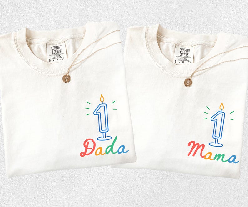 May include: Two off-white t-shirts with the text "Dada" and "Mama" in rainbow colors, each featuring a number one candle graphic. Gold necklaces with round pendants are draped on the shirts. The shirts are from Comfort Colors.