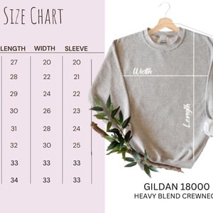 Pode incluir: Uma camisola cinza claro Gildan 18000 Heavy Blend com gola redonda est&aacute; pendurada num cabide de madeira. Uma tabela de tamanhos mostra as medidas em cent&iacute;metros de comprimento, largura e comprimento da manga para os tamanhos S a 5XL.