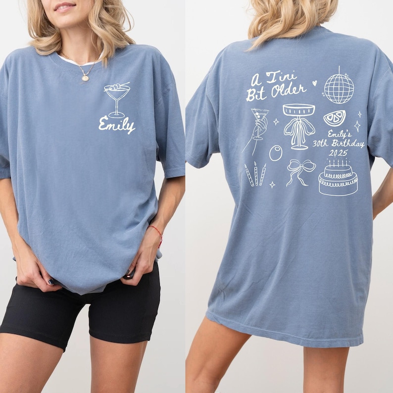 May include: A dusty blue t-shirt with white graphics. The front features a cocktail glass and the name "Emily". The back displays the text "A Tiny Bit Older" with birthday-themed illustrations, including a disco ball and cake. A casual, birthday-themed shirt.