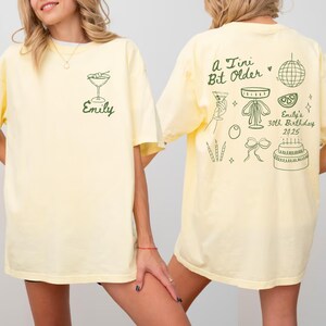 May include: A light yellow t-shirt with a green cocktail graphic and the name "Emily" on the front. The back features a collection of green illustrations, including a disco ball, cake, and text that reads "A Tiny Bit Older" and "Emily's 30th Birthday 2025."